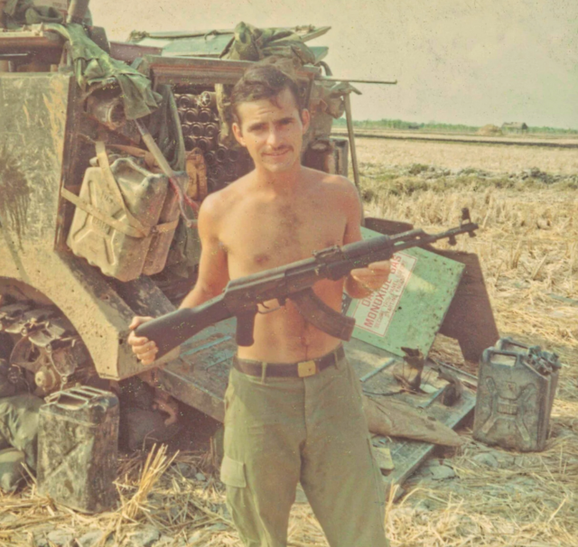 A photo from John’s time in Vietnam. He is standing in front of his armored personnel carrier, holding a captured AK-47 rifle.
